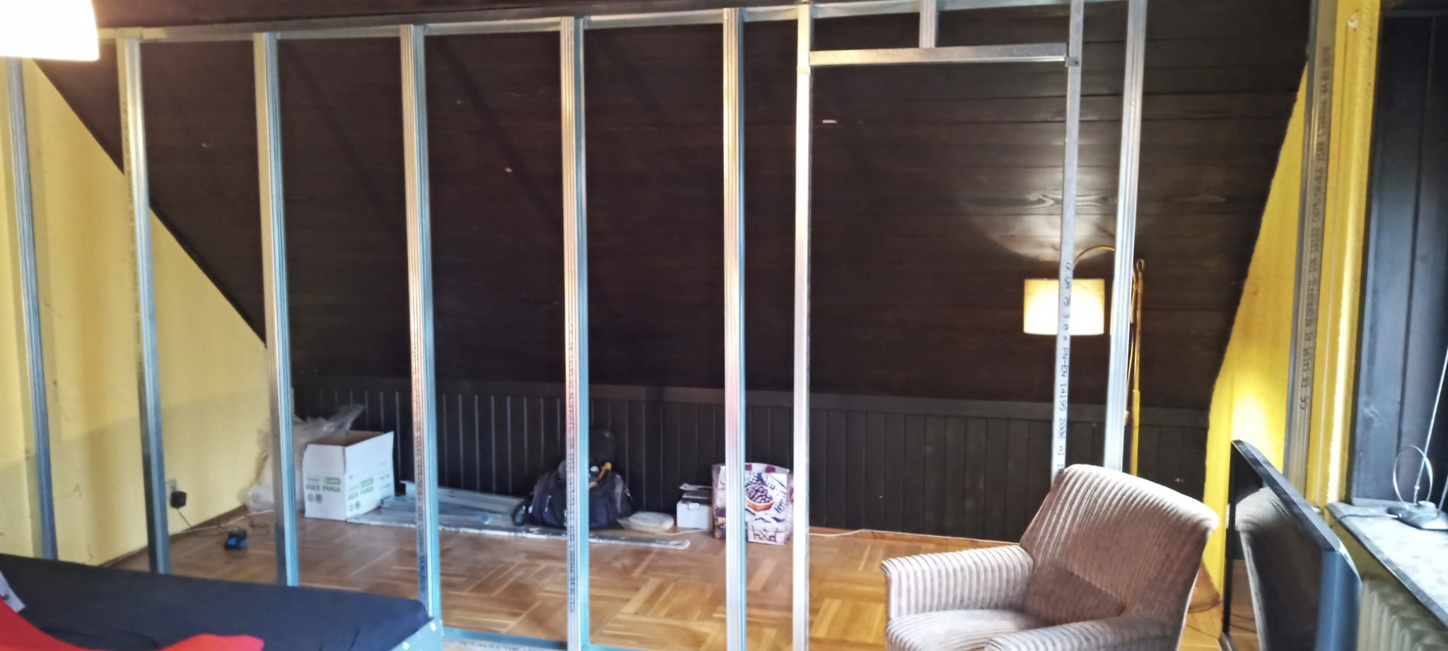 Stelaż z profili metalowych tworzący ścianę działową w pokoju na poddaszu z ukośnym sufitem, widoczne narzędzia i materiały budowlane, w tle ciemna boazeria i lampa podłogowa.