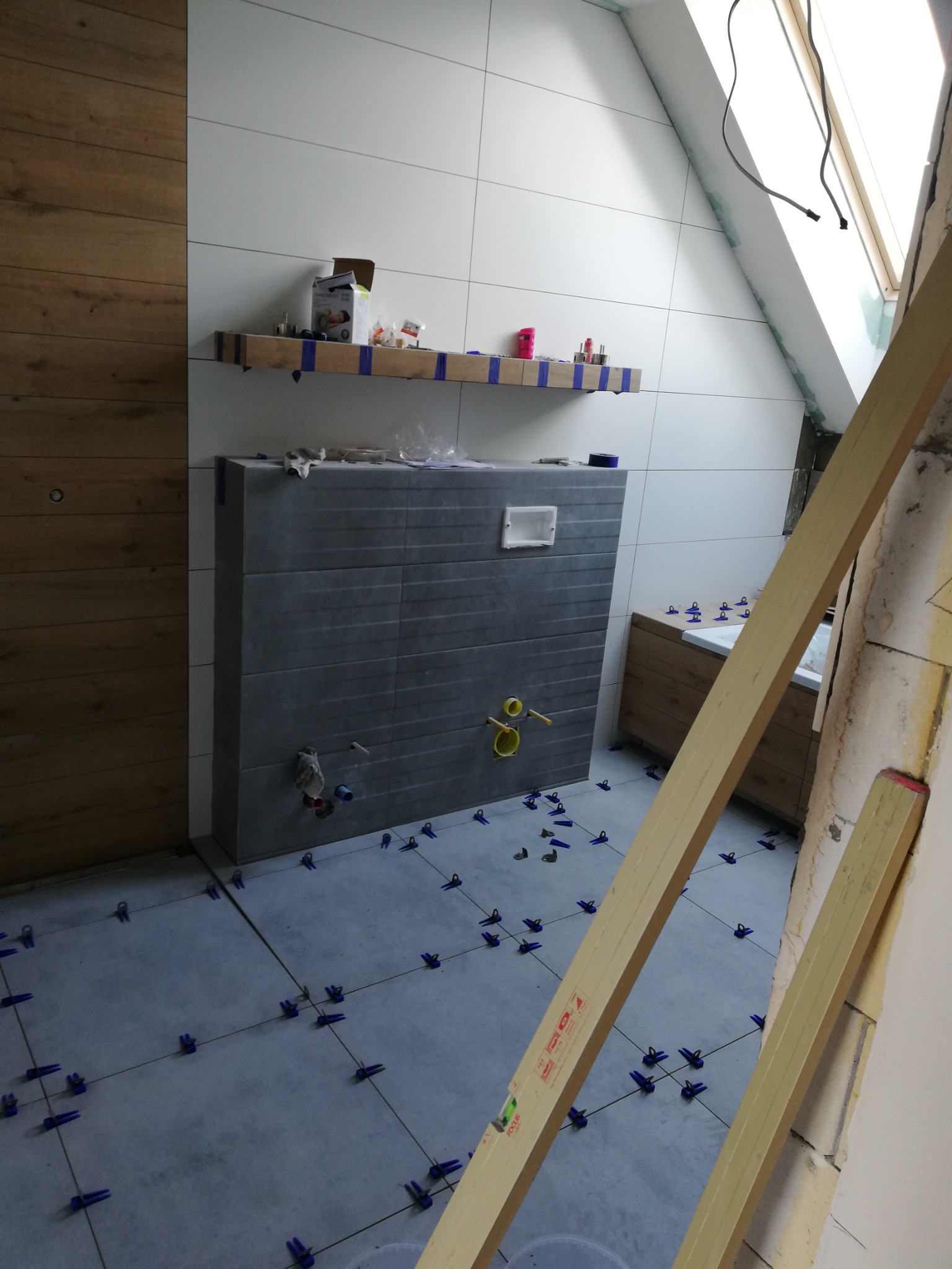 Nowa łazienka w trakcie wykańczania: szare płytki podłogowe z niebieskimi krzyżykami dystansowymi, częściowo wykończone ściany z białych płytek i drewnopodobnej okładziny, widoczny stelaż...