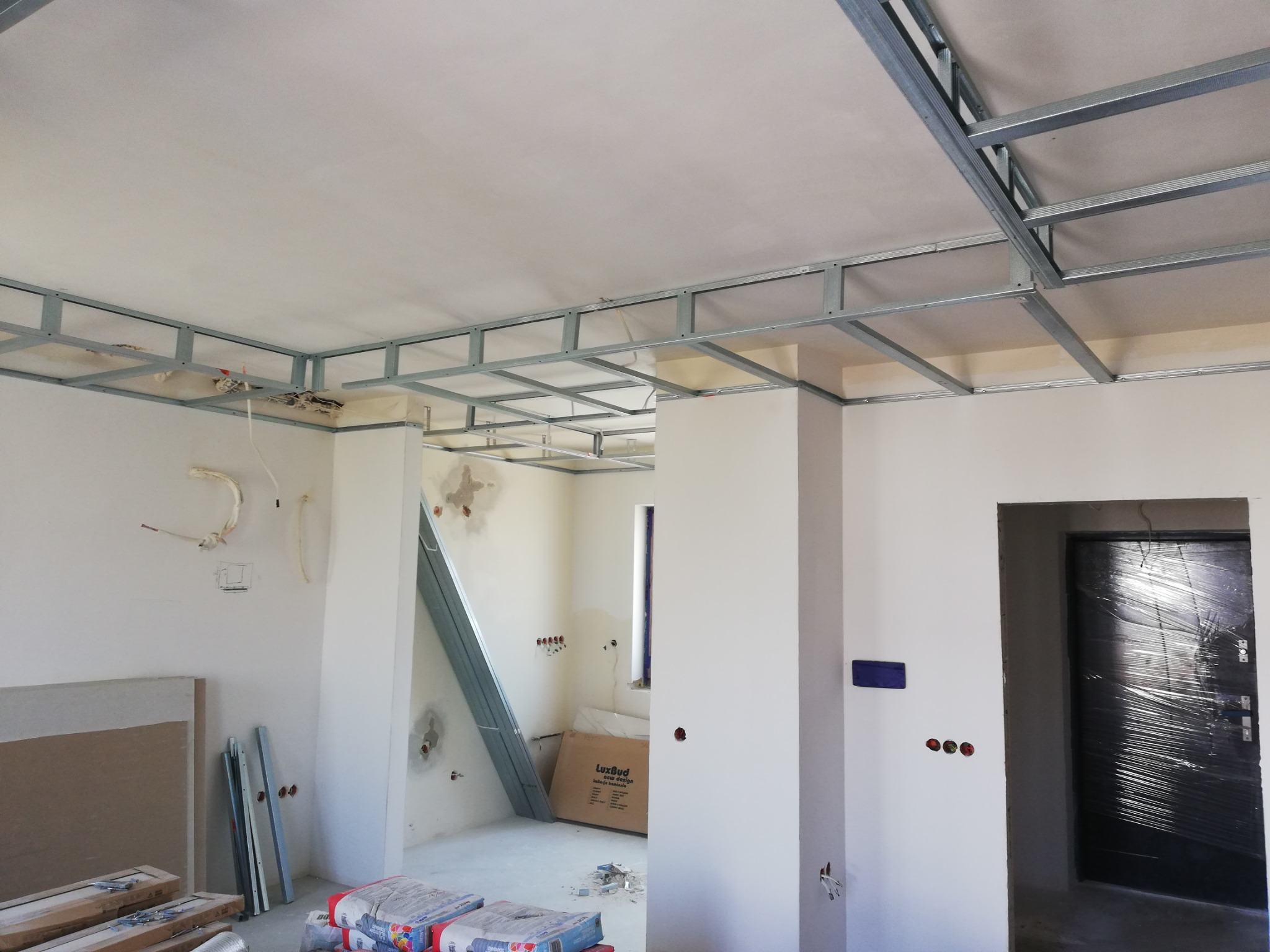 Wnętrze w trakcie remontu: widoczna metalowa konstrukcja sufitu podwieszanego, przygotowane płyty gipsowo-kartonowe i worki z zaprawą, ściany częściowo przygotowane do dalszych prac wykończeniowych...