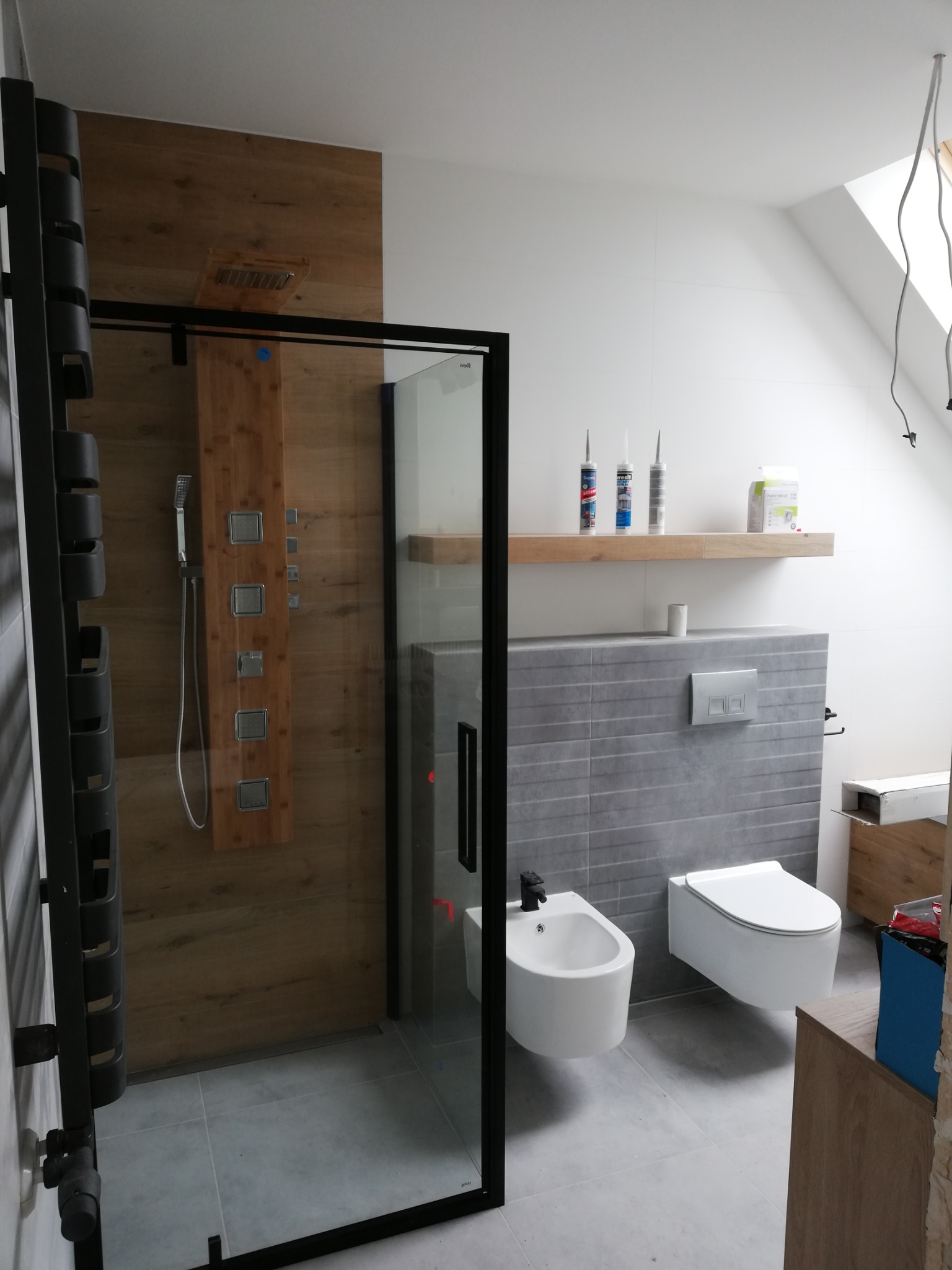 Nowoczesna łazienka z kabiną prysznicową typu walk-in z czarną ramą, drewnopodobną ścianą z panelem prysznicowym, wiszącym bidetem i toaletą, szara płytka na ścianie i podłodze, drewniana półka...