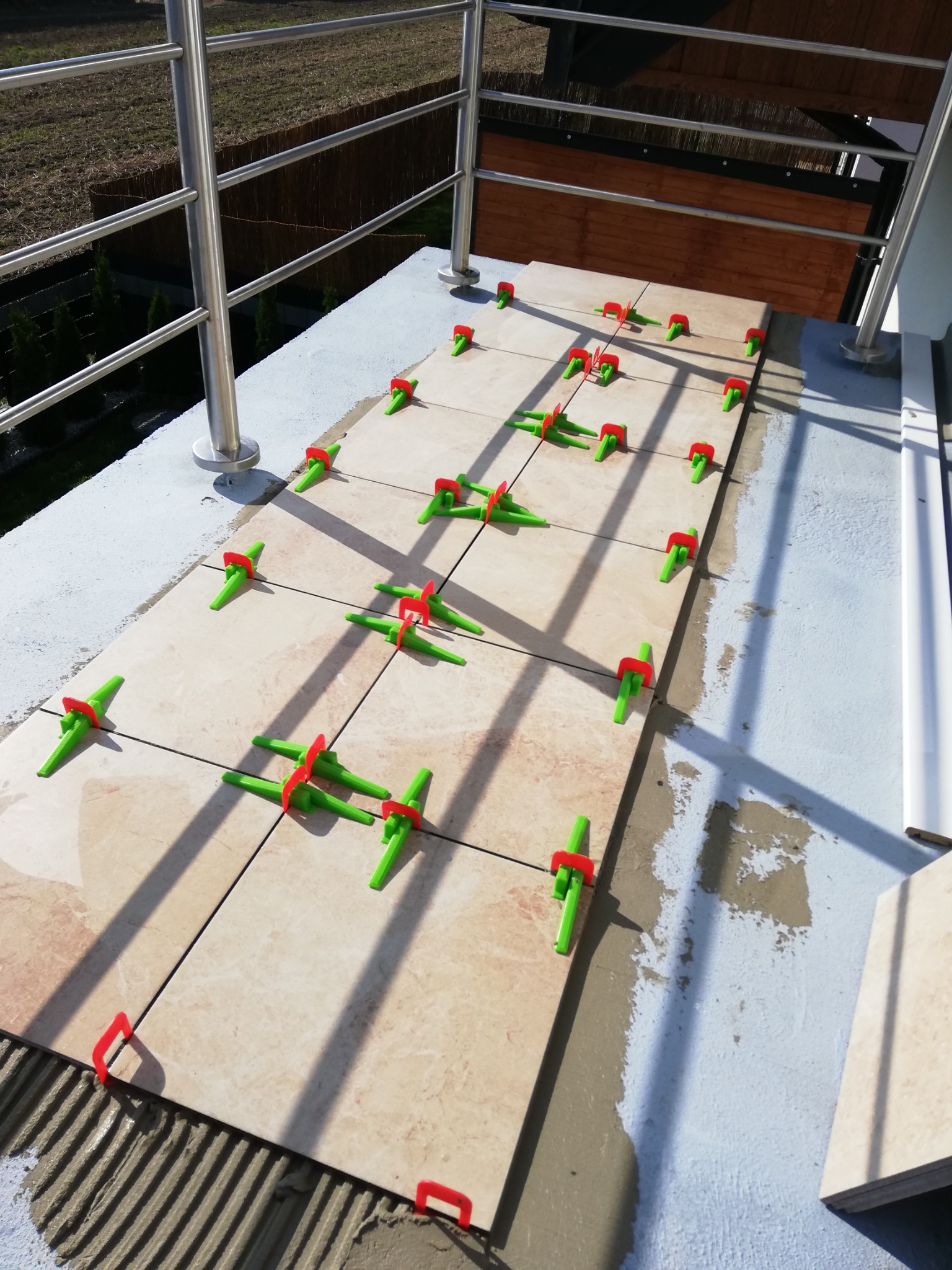 Układanie płytek na balkonie, widoczne płytki z beżowym wzorem, system poziomowania płytek z zielonymi klinami i czerwonymi podstawkami, świeży klej, metalowa balustrada w tle.