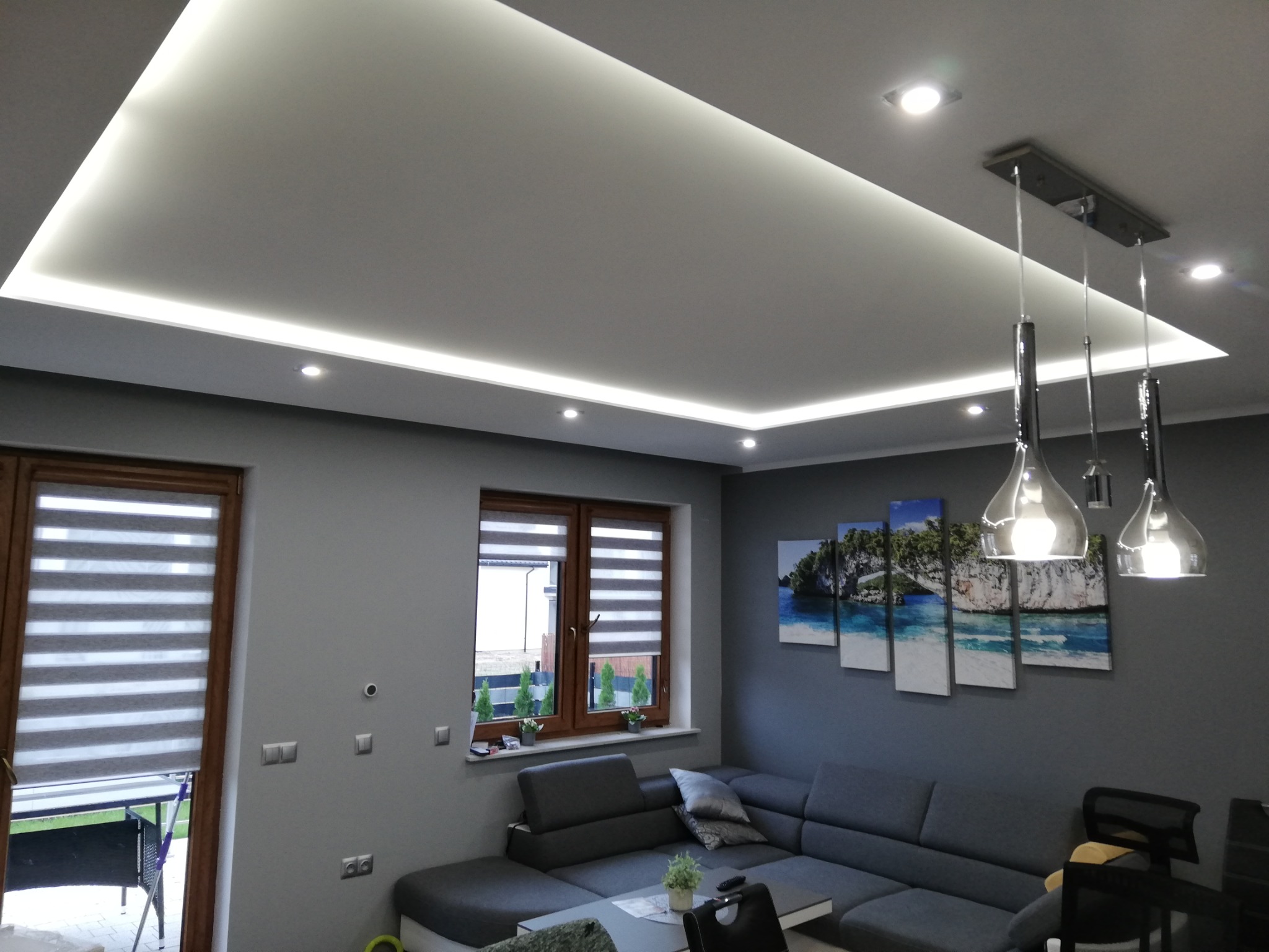 Szare wnętrze salonu z podwieszanym sufitem z oświetleniem LED, nowoczesną lampą wiszącą i szarą sofą narożną.