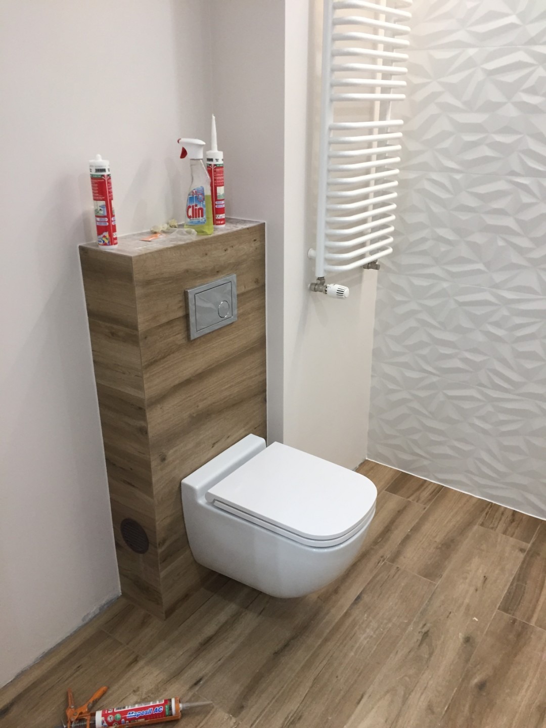 Wisząca toaleta z zabudową w odcieniach drewna, biały grzejnik drabinkowy i trójwymiarowa struktura płytek na ścianie, widoczne materiały budowlane na zabudowie i podłodze.