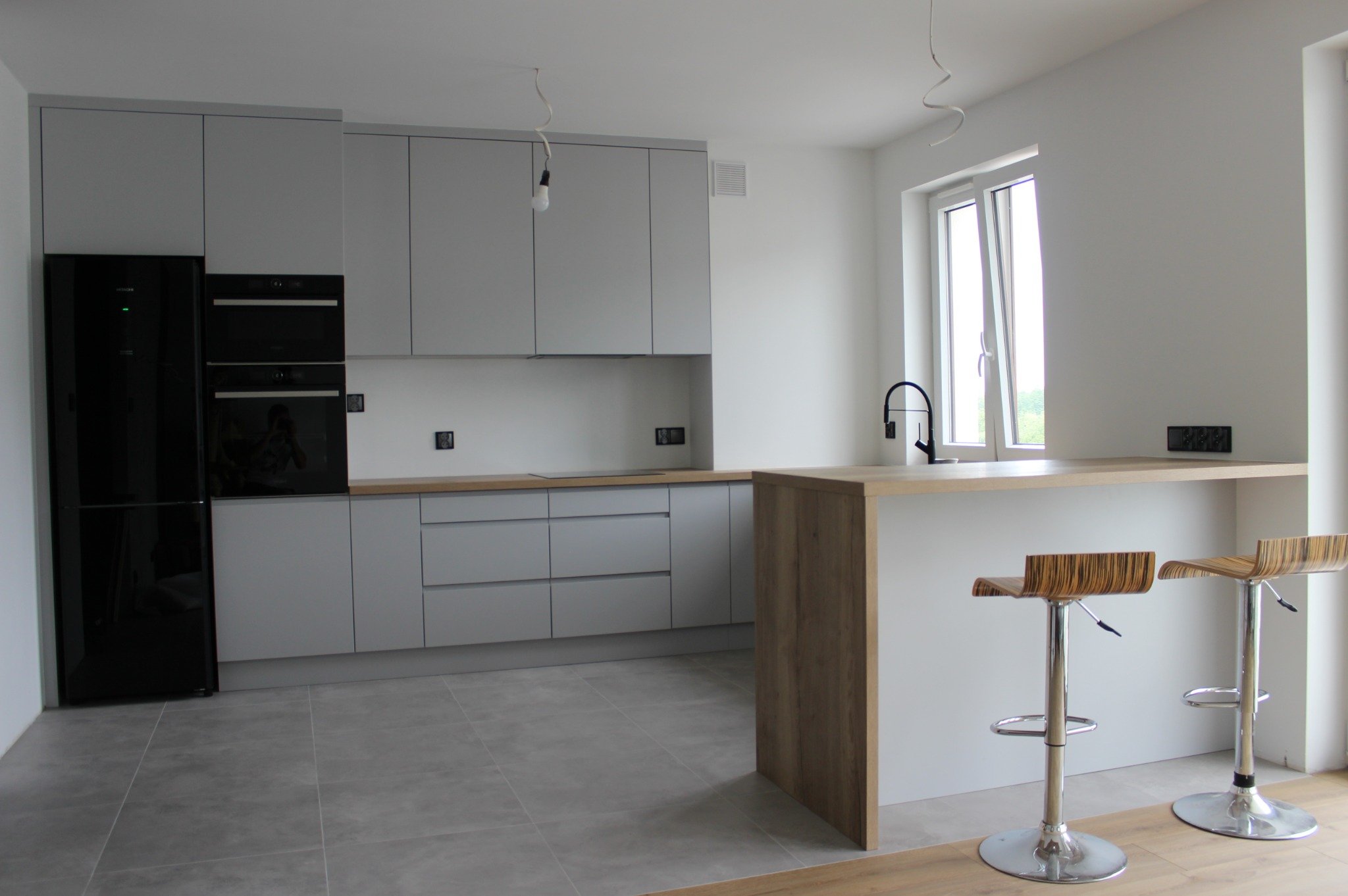 Nowoczesna, minimalistyczna kuchnia w odcieniach szarości z drewnianym blatem i barkiem. Dwa metalowe hokery z drewnianymi siedziskami stoją przy barku. Na podłodze szare płytki.