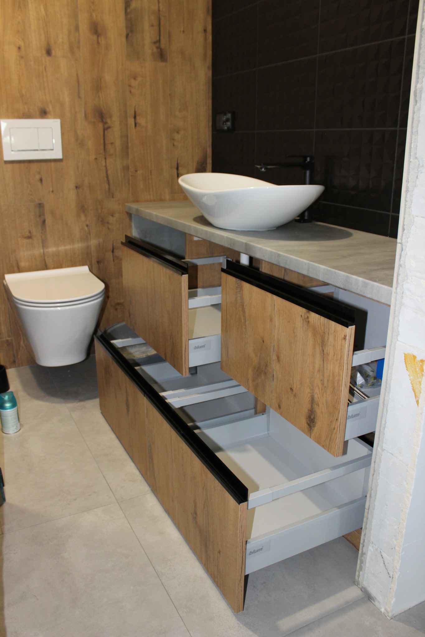 Łazienka z drewnianą zabudową umywalki z szufladami Blum, białą umywalką nablatową i toaletą podwieszaną na tle drewnianej i czarnej ściany.