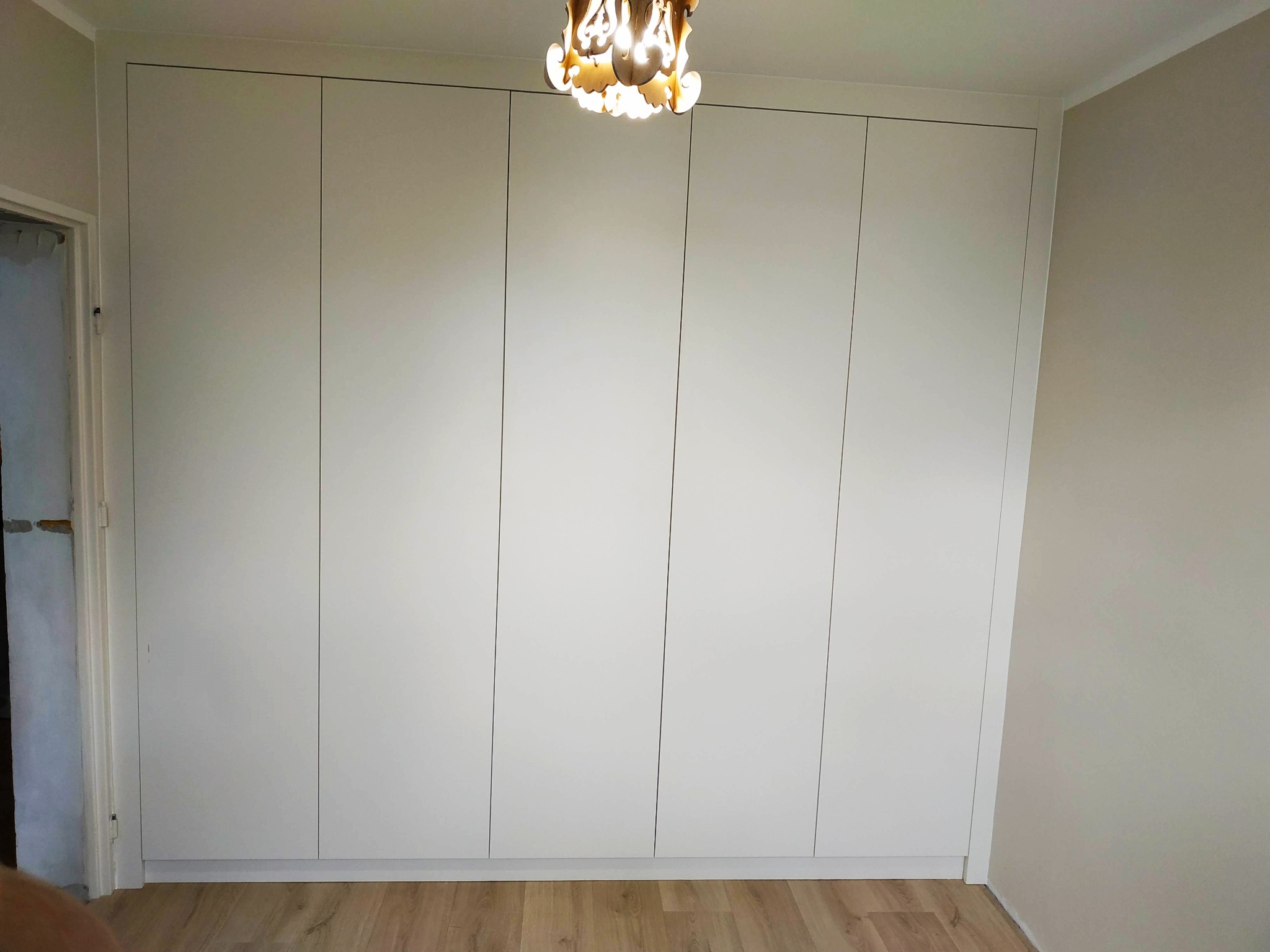 Biała, minimalistyczna szafa wnękowa z pięcioma drzwiami, dopasowana do sufitu, z widocznym drewnianym parkietem i ozdobną lampą na suficie.