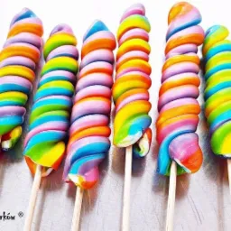 Siedem tęczowych lizaków w kształcie spirali na drewnianych patyczkach, ułożonych obok siebie na metalowej powierzchni, z widocznym logo 'Manufaktura Cukierków' w lewym dolnym rogu.