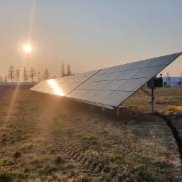 Instalacja paneli słonecznych na łące w promieniach wschodzącego słońca, widoczny wykop pod kable i ślady opon po ciężkim sprzęcie.