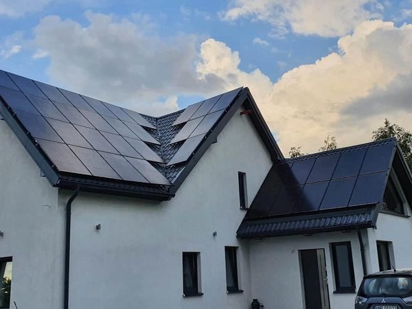 Czarny system paneli słonecznych na dachu dwuspadowym nowoczesnego domu z białym tynkiem, widok z dołu na tle błękitnego nieba z chmurami.