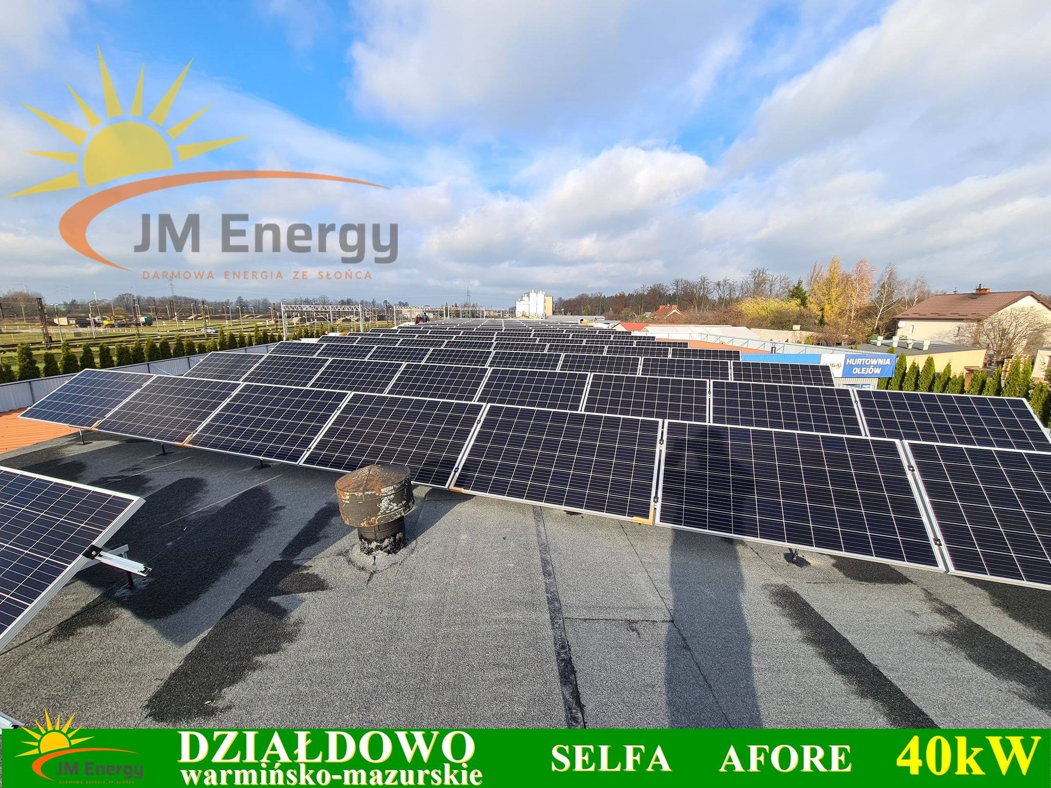 Rozległa instalacja paneli słonecznych na płaskim dachu z widocznym kominem wentylacyjnym, logo firmy JM Energy i napisy Działdowo, warmińsko-mazurskie, SELFA, AFORE, 40kW. W tle budynki i błękitne...
