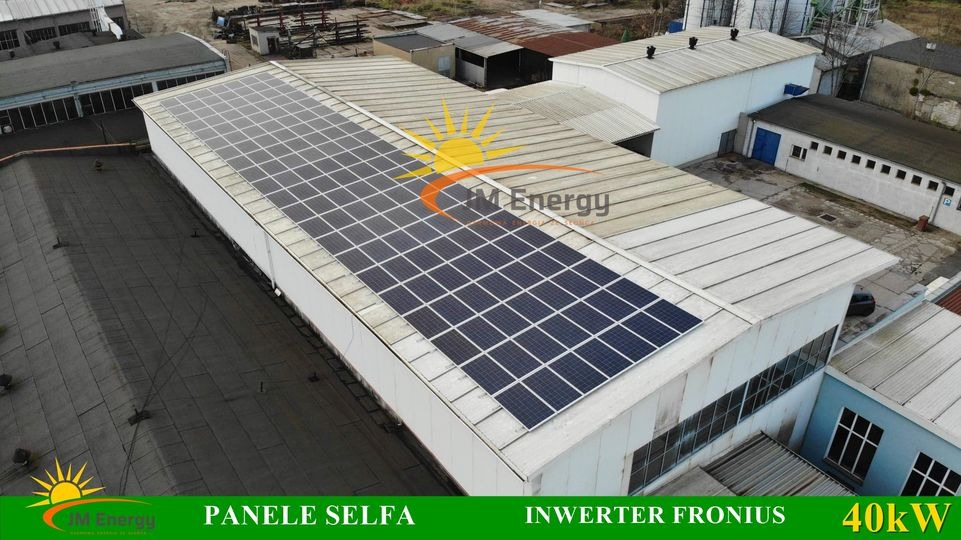 Widok z góry na instalację paneli słonecznych na dachu hali przemysłowej; widoczne logo firmy JM Energy, napisy 'Panele Selfa' i 'Inverter Fronius 40kW'.