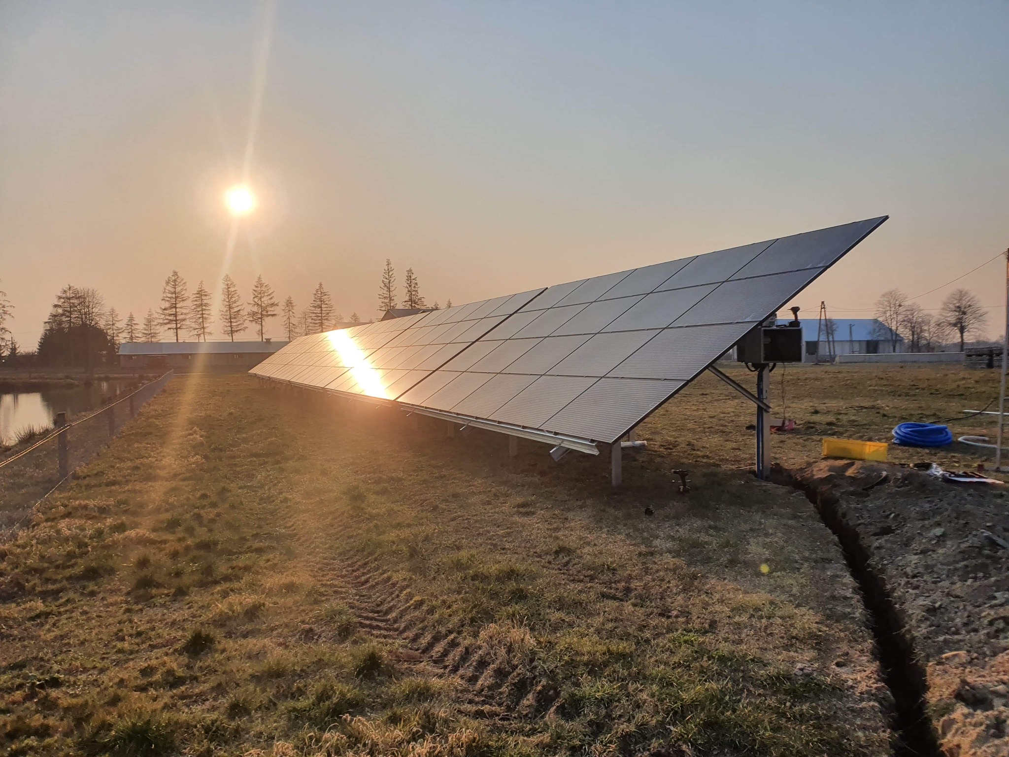 Instalacja paneli słonecznych na łące w promieniach wschodzącego słońca, widoczny wykop pod kable i ślady opon po ciężkim sprzęcie.