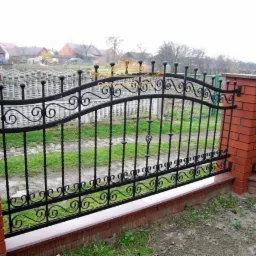 Kute ogrodzenie z czarnego metalu z dekoracyjnymi elementami i ceglanymi słupkami, widoczne z bliska na tle zabudowań.