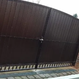 Ciemnobrązowa brama wjazdowa dwuskrzydłowa z metalowymi panelami i ozdobnym dolnym wykończeniem, osadzona na ceglanych słupkach, z logiem firmy w prawym górnym rogu.