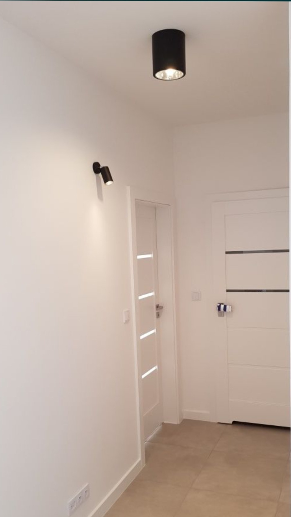 Białe wnętrze korytarza z dwiema białymi parami drzwi, oświetlone czarną lampą sufitową i reflektorem ściennym.