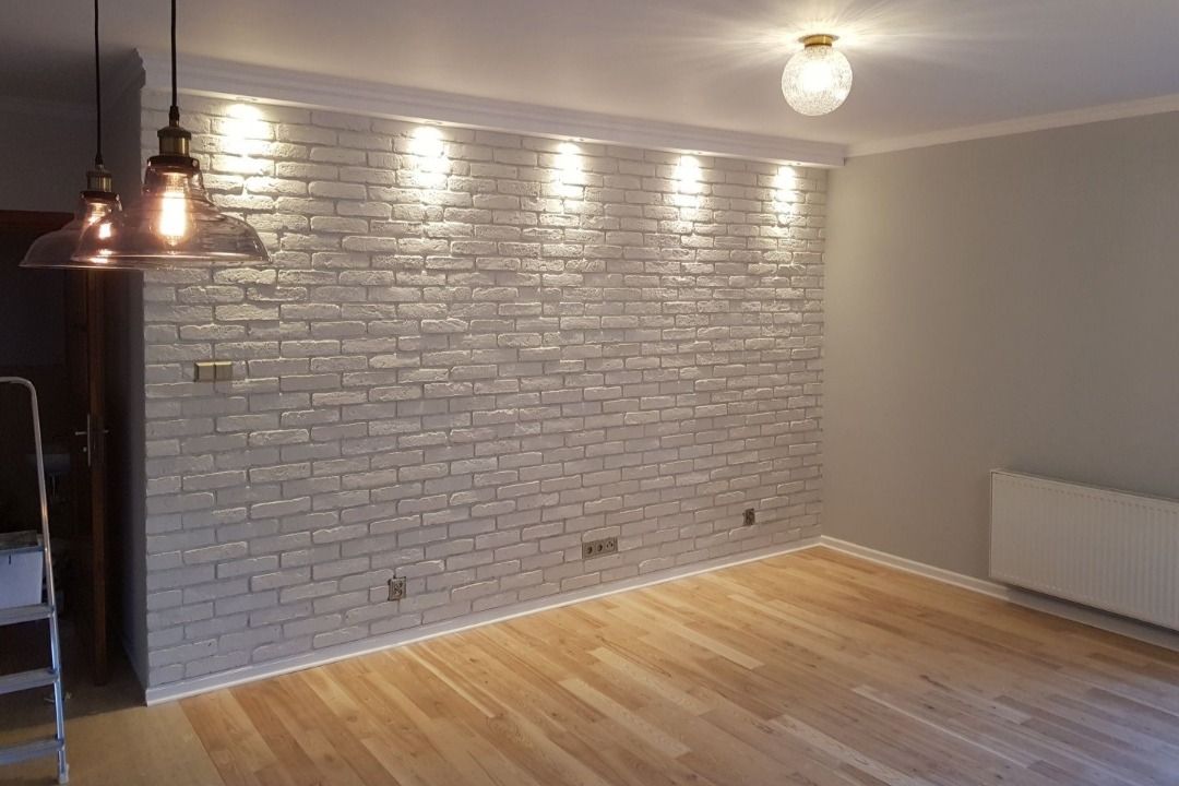 Pokój po remoncie z białą ścianą z cegły, oświetleniem punktowym i drewnianą podłogą; widoczne wiszące lampy w stylu industrialnym i drabina malarska z boku.