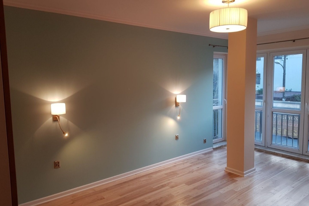 Świeżo odmalowane wnętrze w Warszawie z drewnianą podłogą, ścianami w odcieniach zieleni i nowoczesnymi kinkietami.