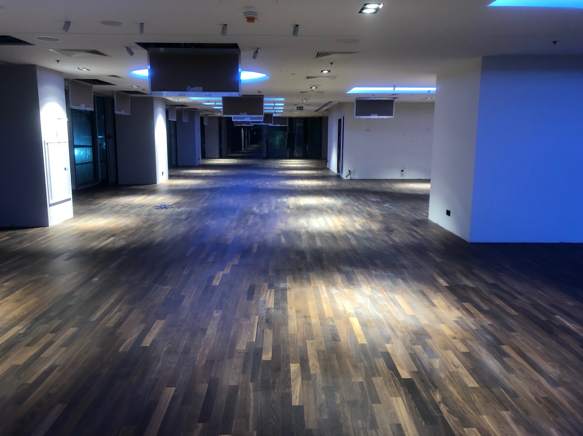 Długi korytarz z ciemną, drewnianą podłogą ułożoną w jodełkę. Sufit z podwieszanymi elementami i oświetleniem LED. Widok w głąb korytarza, perspektywa zbieżna.