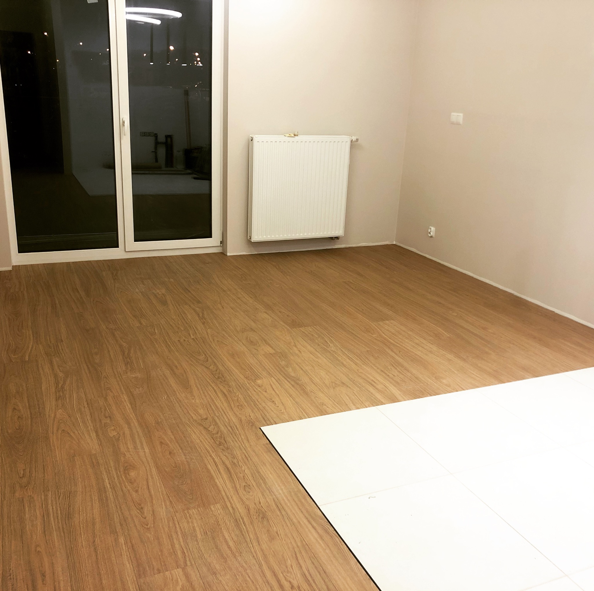 Jasne panele podłogowe imitujące drewno w pokoju w Warszawie, z fragmentem białych płytek. Widoczny grzejnik i okno balkonowe.