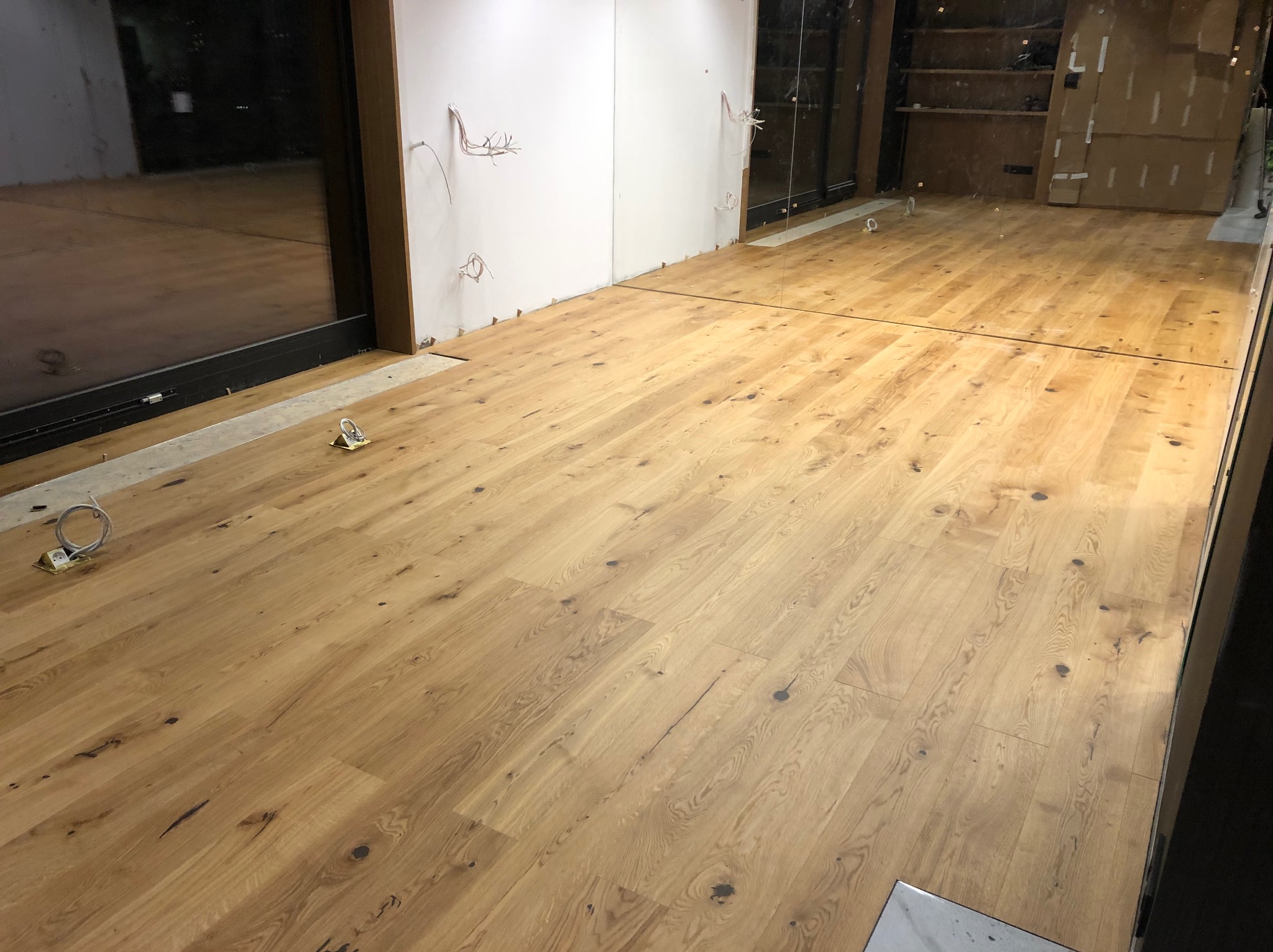 Jasny, dębowy parkiet ułożony w nowym wnętrzu z widocznymi przewodami elektrycznymi na ścianach. Minimalistyczny design, surowe wykończenie, ciepłe odcienie drewna.