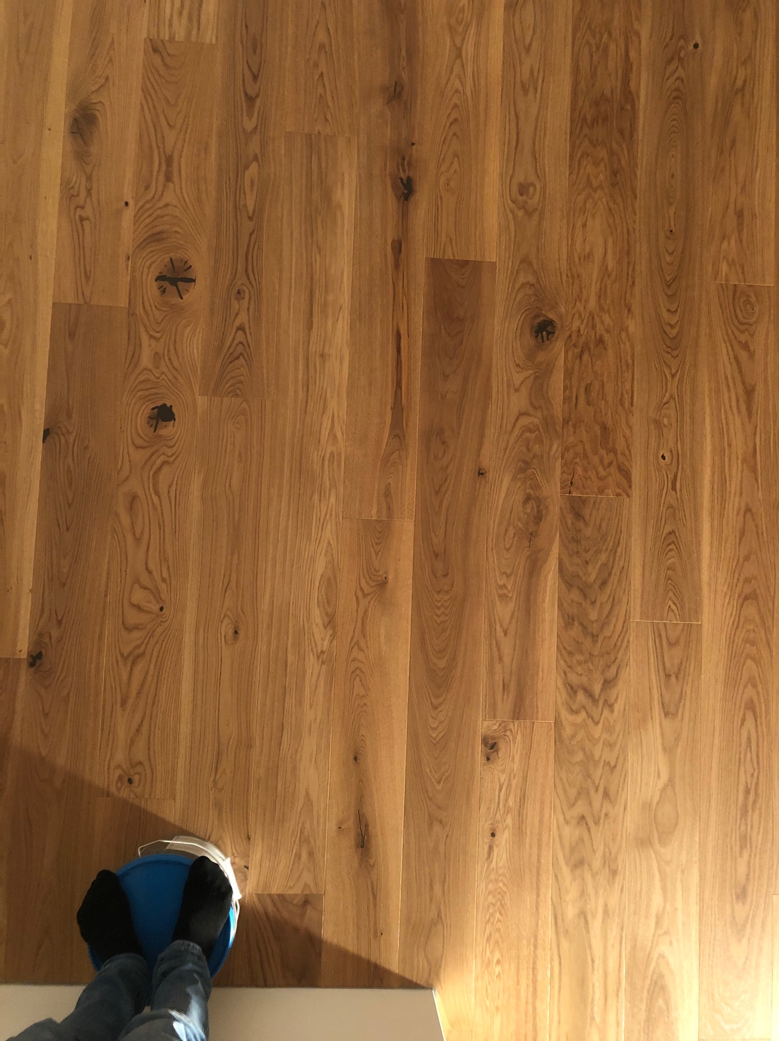 Ułożony parkiet z naturalnego drewna z widocznymi słojami i sękami, na którym stoi osoba w skarpetkach na niebieskim wiadrze. Widok z góry, ciepłe oświetlenie.