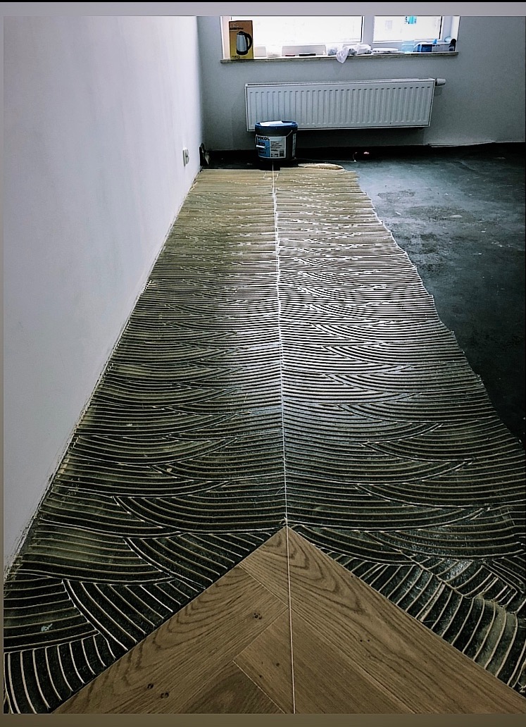 Fragment układania parkietu w jodełkę. Widoczny klej na podłodze i ułożone deski. W tle wiadro z klejem i grzejnik pod oknem. Ujęcie z niskiej perspektywy.