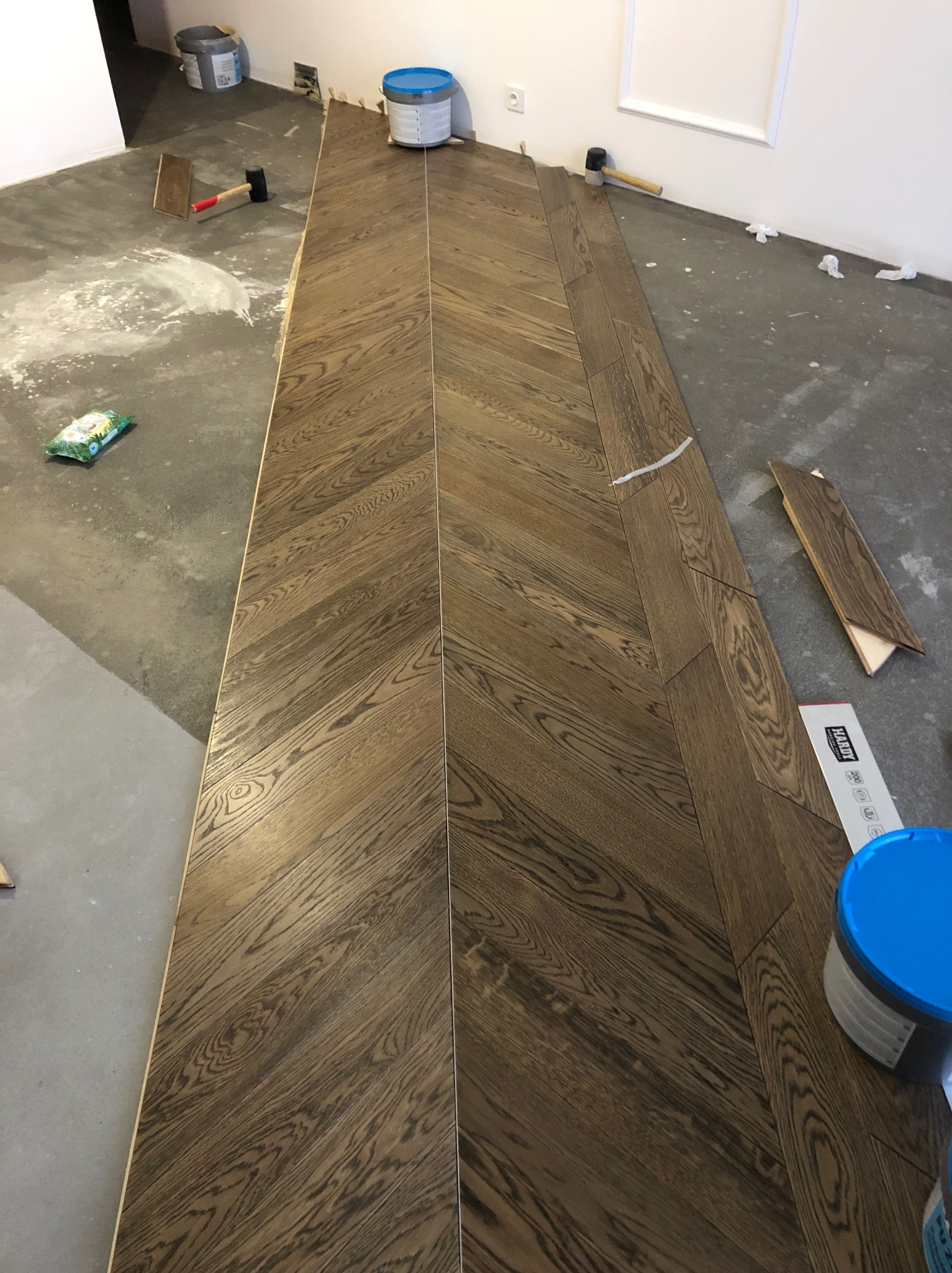 Układanie parkietu w jodełkę. Widoczne narzędzia i materiały: młotek, klej, listwy. Częściowo ułożona podłoga z ciemnego drewna na szarym podłożu.