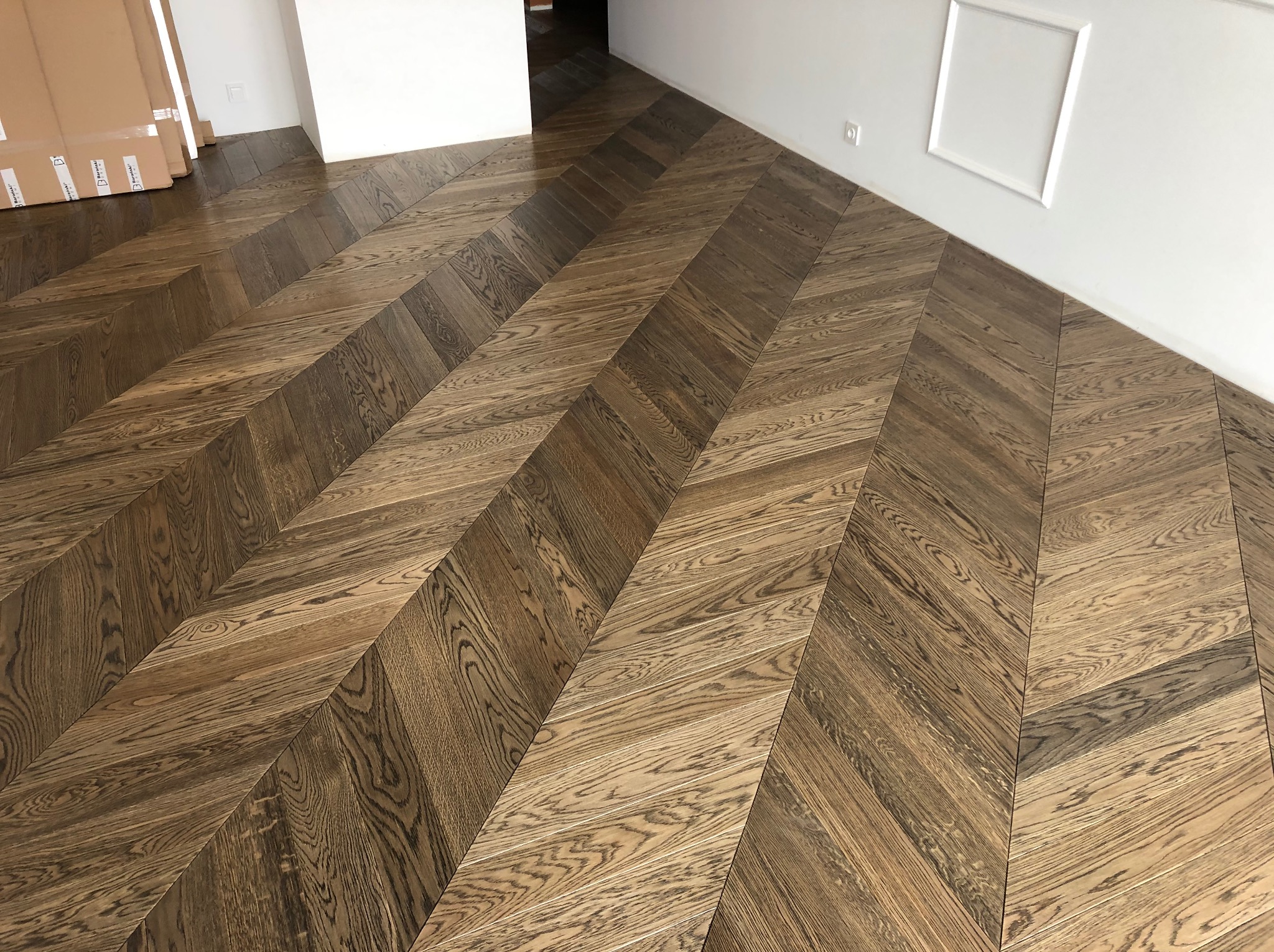 Ukończona podłoga z parkietu ułożonego w jodełkę, z widocznymi paczkami parkietu przy ścianie. Białe ściany z dekoracyjną listwą.