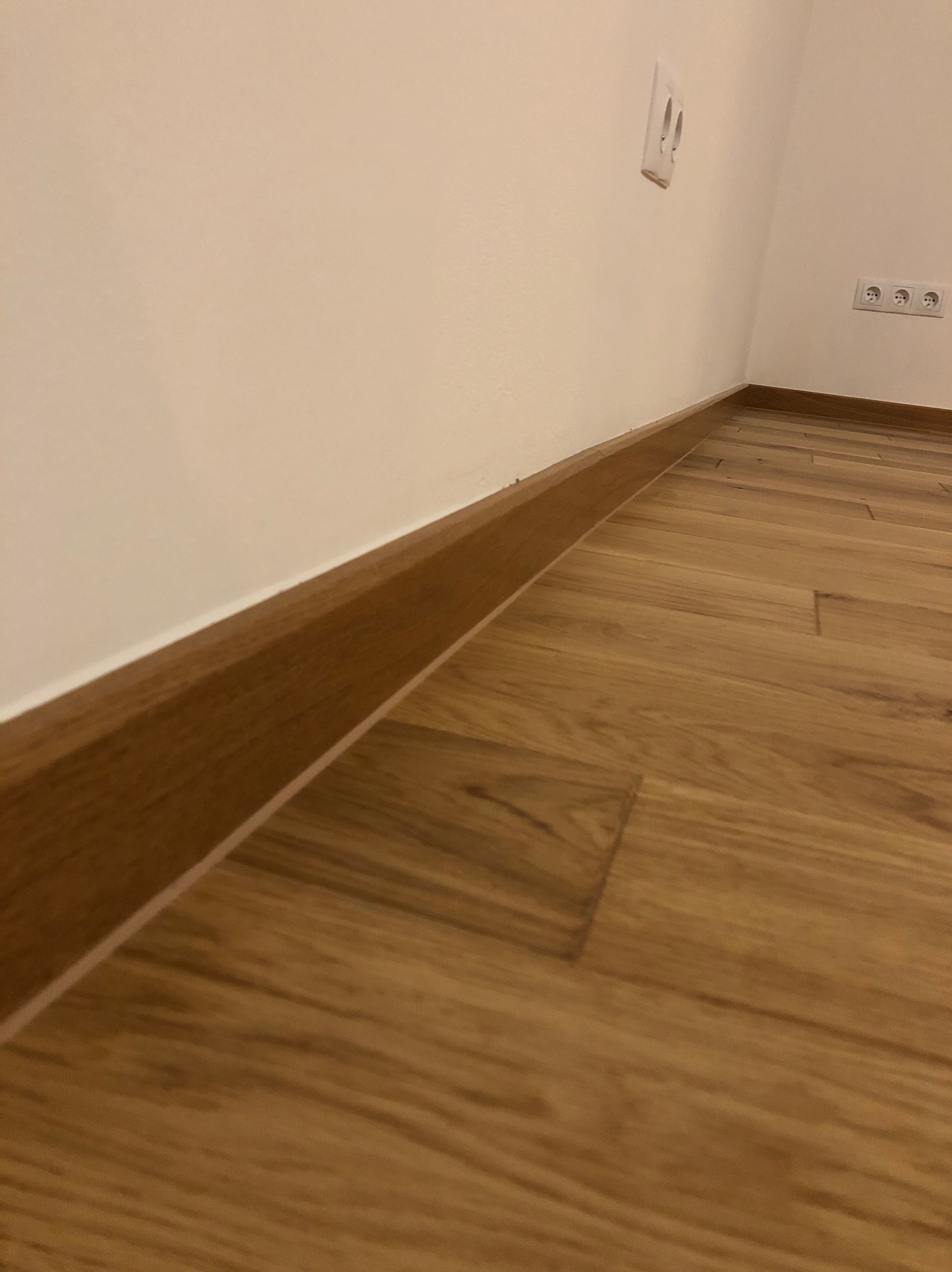 Ukończona podłoga z paneli drewnopodobnych z brązową listwą przypodłogową przy białej ścianie z gniazdkami elektrycznymi. Ujęcie z niskiej perspektywy.