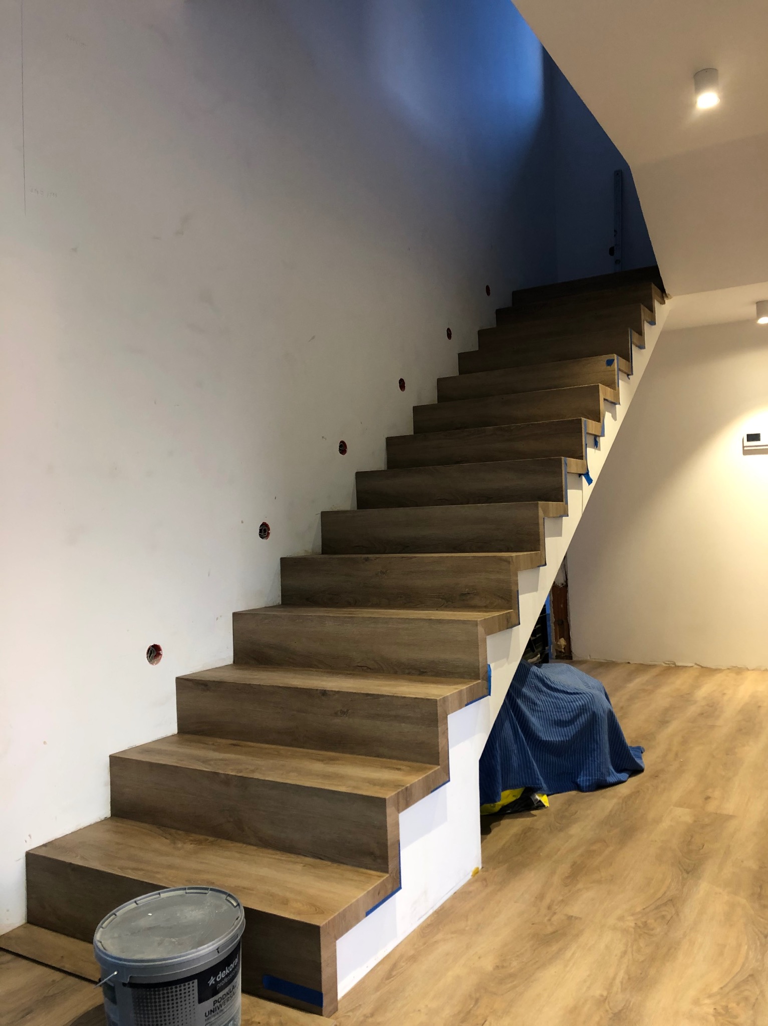 Schody wykończone panelami drewnopodobnymi, z widocznym wiadrem farby i zabezpieczeniem. Ściany białe, przygotowane do malowania. Oświetlenie sufitowe.