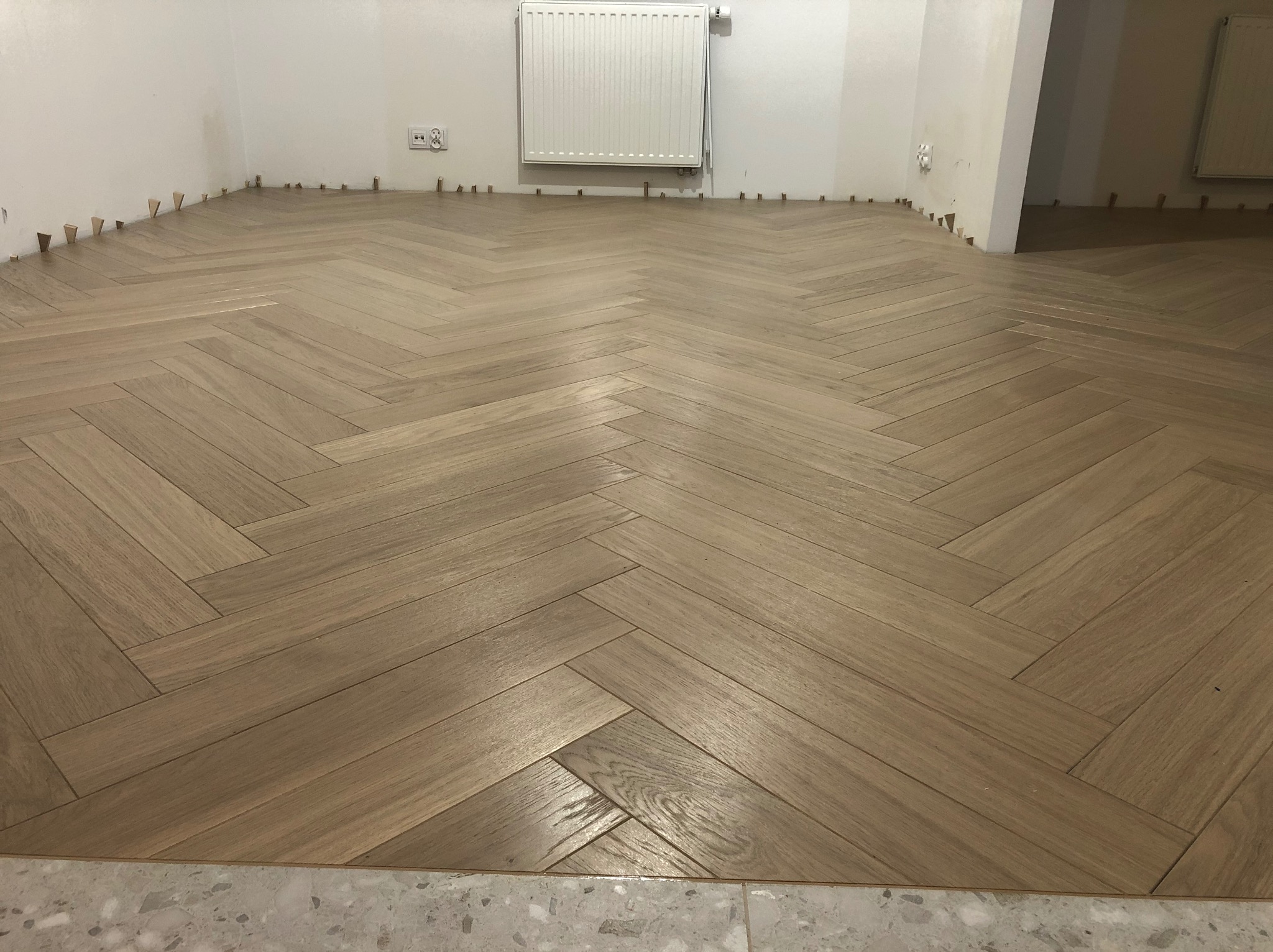 Ułożony parkiet w jodełkę, jasne drewno, widoczne kliny dystansowe przy ścianach, grzejnik na ścianie. Fragment podłogi z płytek w dolnej części kadru.