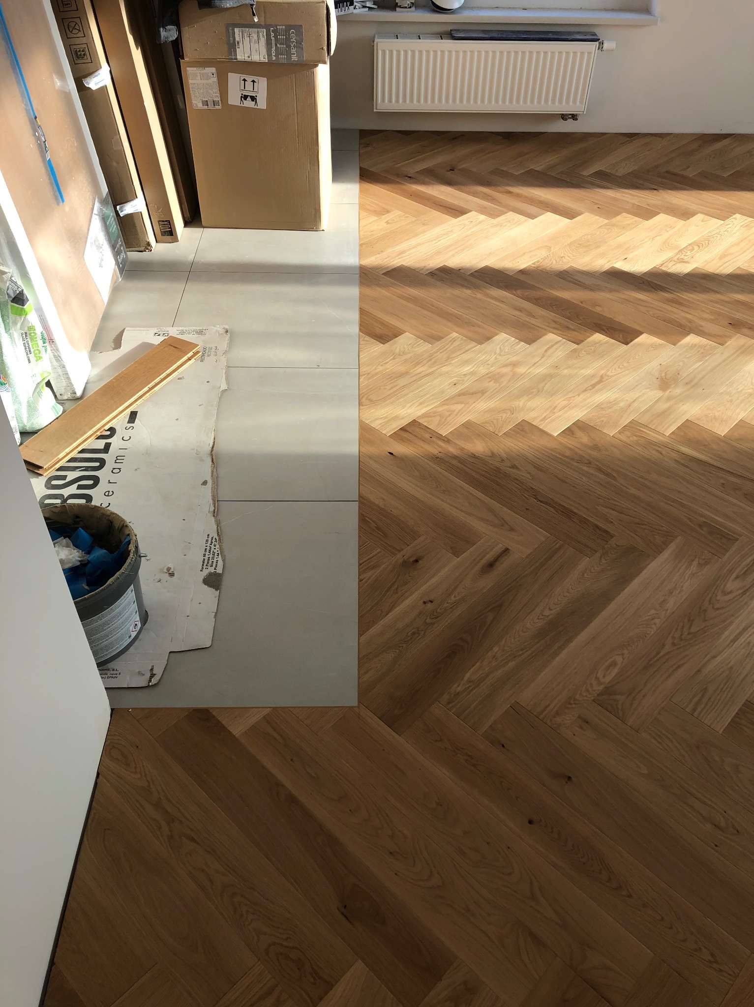 Fragment podłogi z ułożonym parkietem w jodełkę, kontrastujący z szarymi płytkami i materiałami budowlanymi. Widoczny grzejnik i kartony z płytkami.
