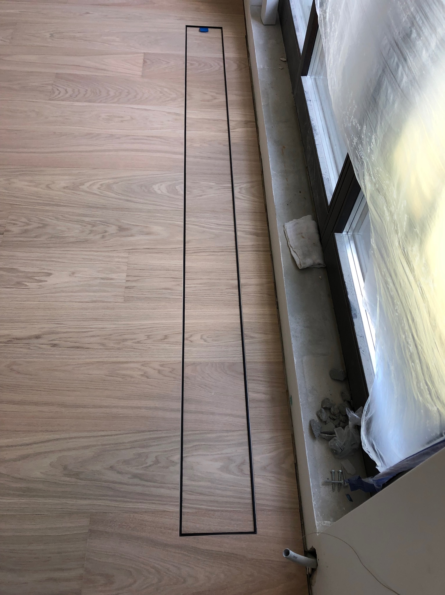 Jasne panele podłogowe z wąskim, czarnym odpływem liniowym wzdłuż okna zabezpieczonego folią. Widoczne drobne elementy montażowe i fragment instalacji.