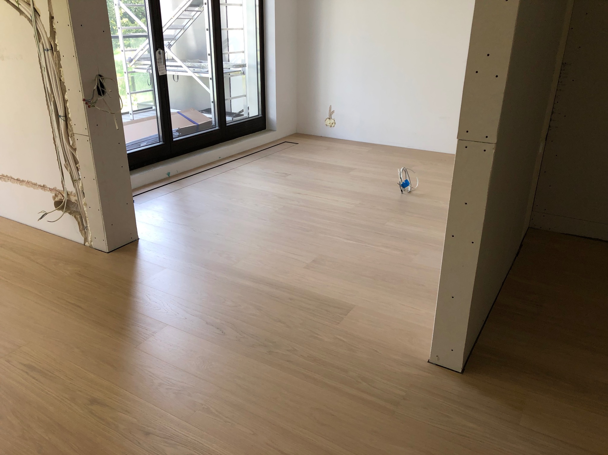 Jasne panele podłogowe w pokoju z widoczną instalacją elektryczną w ścianach i czarną listwą przypodłogową. Widok na okno balkonowe i rusztowanie na zewnątrz.