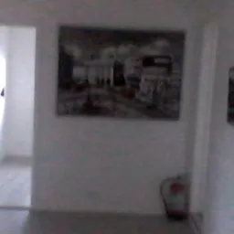 Jasny korytarz z białymi ścianami, ozdobiony obrazami i wyposażony w gaśnicę przy podłodze.