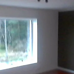 Pomalowane na biało ściany pokoju z dużym oknem, przez które widać zalesiony teren i mały czerwony budynek w tle; jedna ściana pomalowana na ciemny kolor.