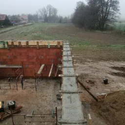 Wznoszenie muru z czerwonej cegły i betonowanie fundamentów na placu budowy w pochmurny dzień. Widoczne szalunki, rusztowania i narzędzia.