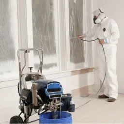 Mężczyzna w kombinezonie ochronnym maluje okno natryskowo, widoczny agregat malarski i folia zabezpieczająca.