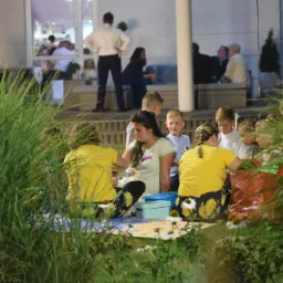 Dwie kobiety w żółtych koszulkach i dzieci siedzą na kocu na trawie, być może podczas animacji na świeżym powietrzu, w tle widać budynek i osoby.