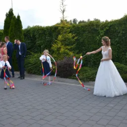 Panna młoda w białej sukni trzyma kolorowe wstążki, a dwie dziewczynki w granatowych sukienkach również bawią się wstążkami na tle zielonego żywopłotu i innych gości weselnych.