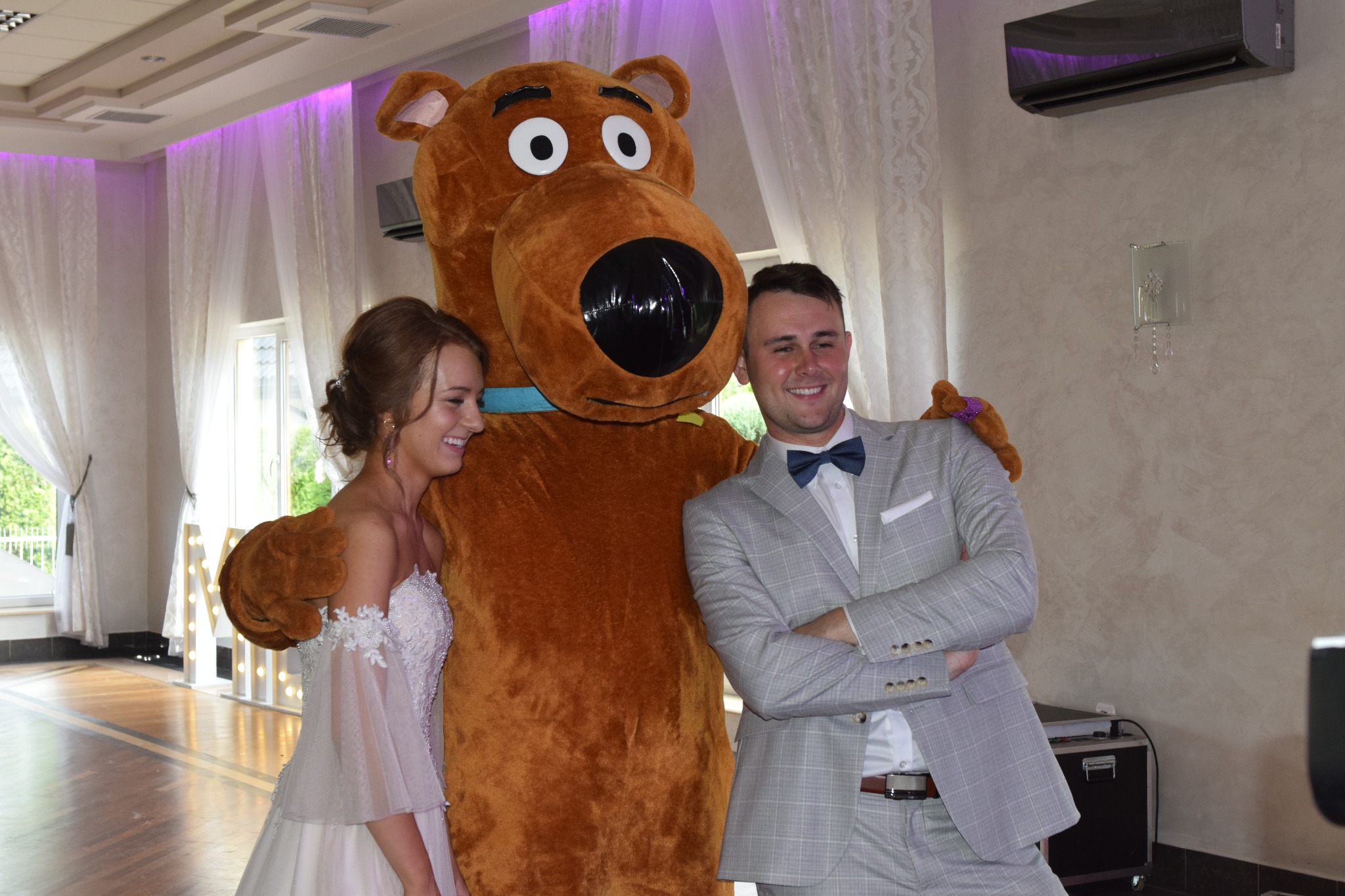 Uśmiechnięta panna młoda w białej sukni i pan młody w szarym garniturze z muszką pozują z dużą maskotką brązowego psa w sali z oknami i białymi zasłonami.