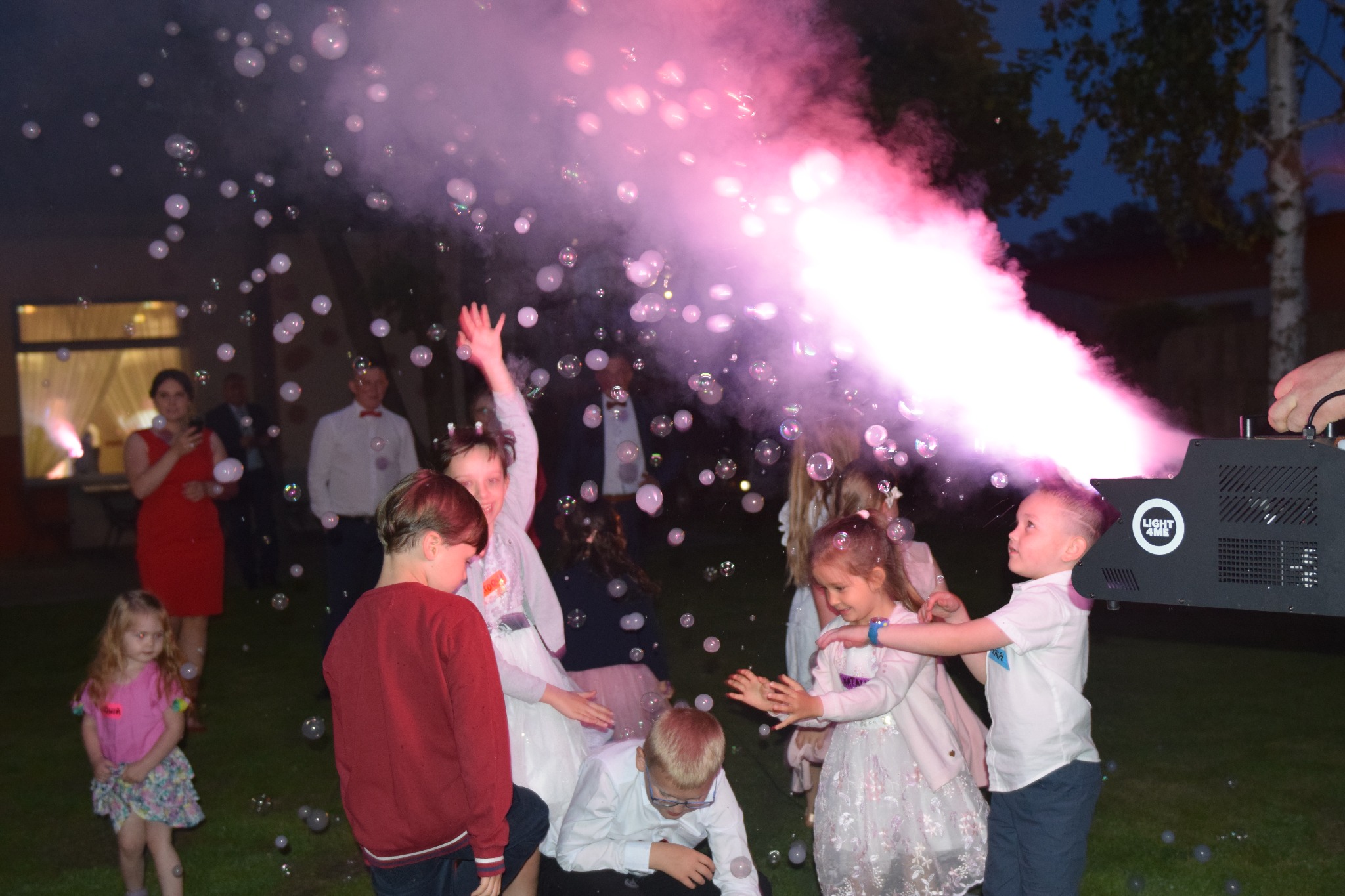 Grupa dzieci bawi się w otoczeniu baniek mydlanych, które są wypuszczane z profesjonalnej maszyny do baniek z logo 'LIGHT 4ME', w tle widoczni dorośli i budynek.