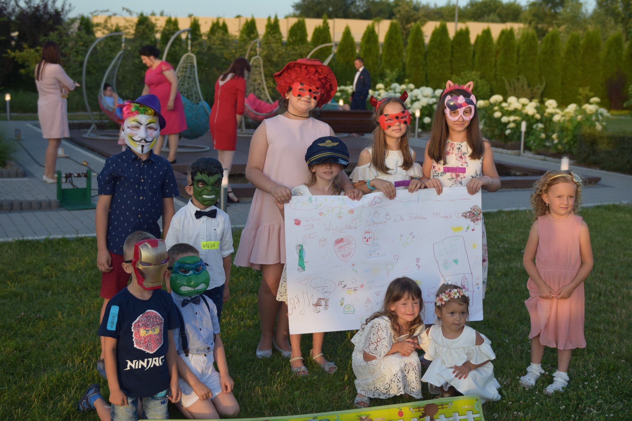 Grupa dzieci w maskach superbohaterów i ozdobnych nakryciach głowy pozuje z dużym rysunkiem na papierze, prawdopodobnie prezentem dla młodej pary, w tle widoczne osoby dorosłe i huśtawki ogrodowe.
