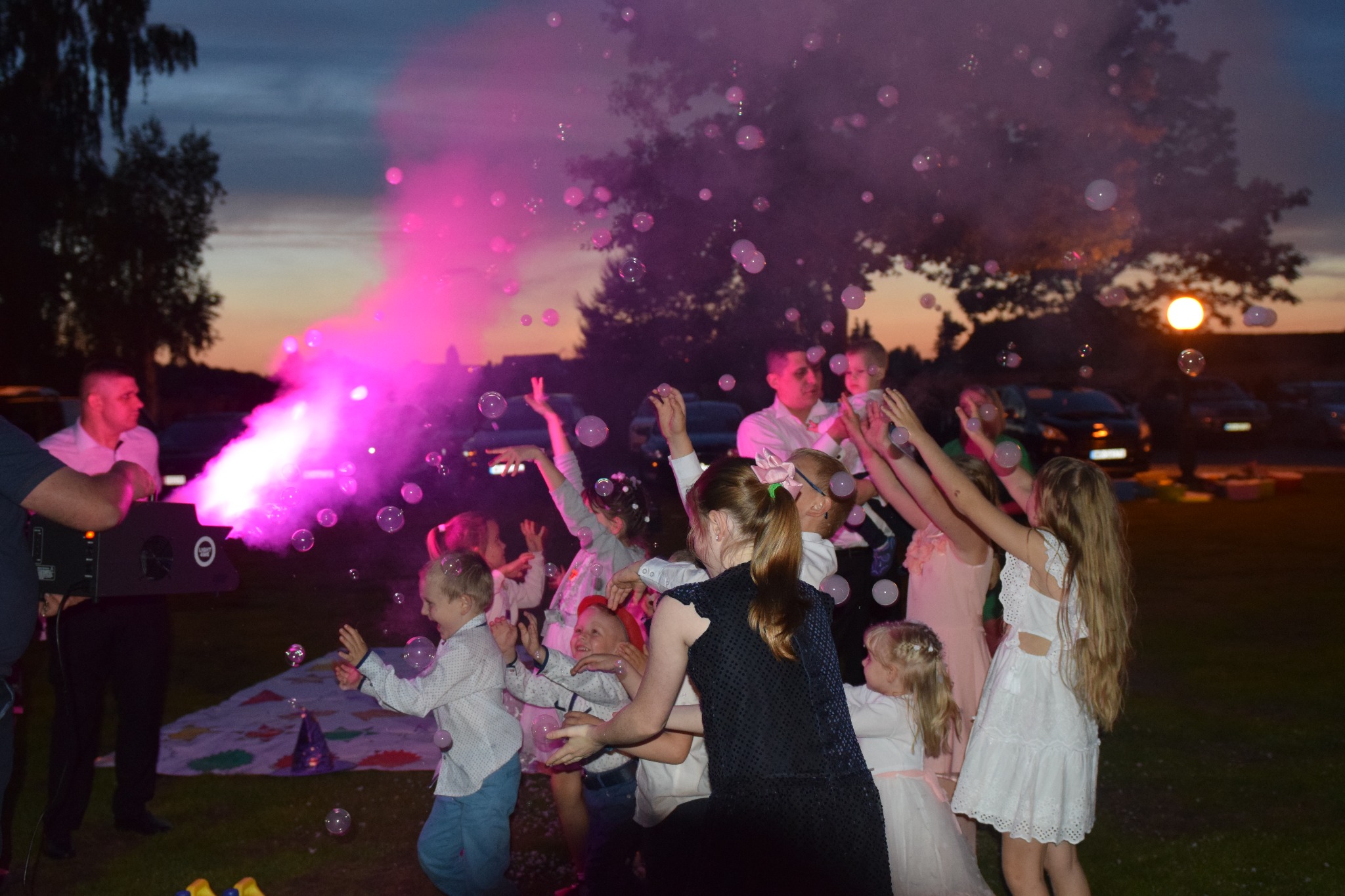 Grupa dzieci bawi się w otoczeniu baniek mydlanych wytwarzanych przez maszynę z różowym oświetleniem podczas wieczornej imprezy plenerowej.