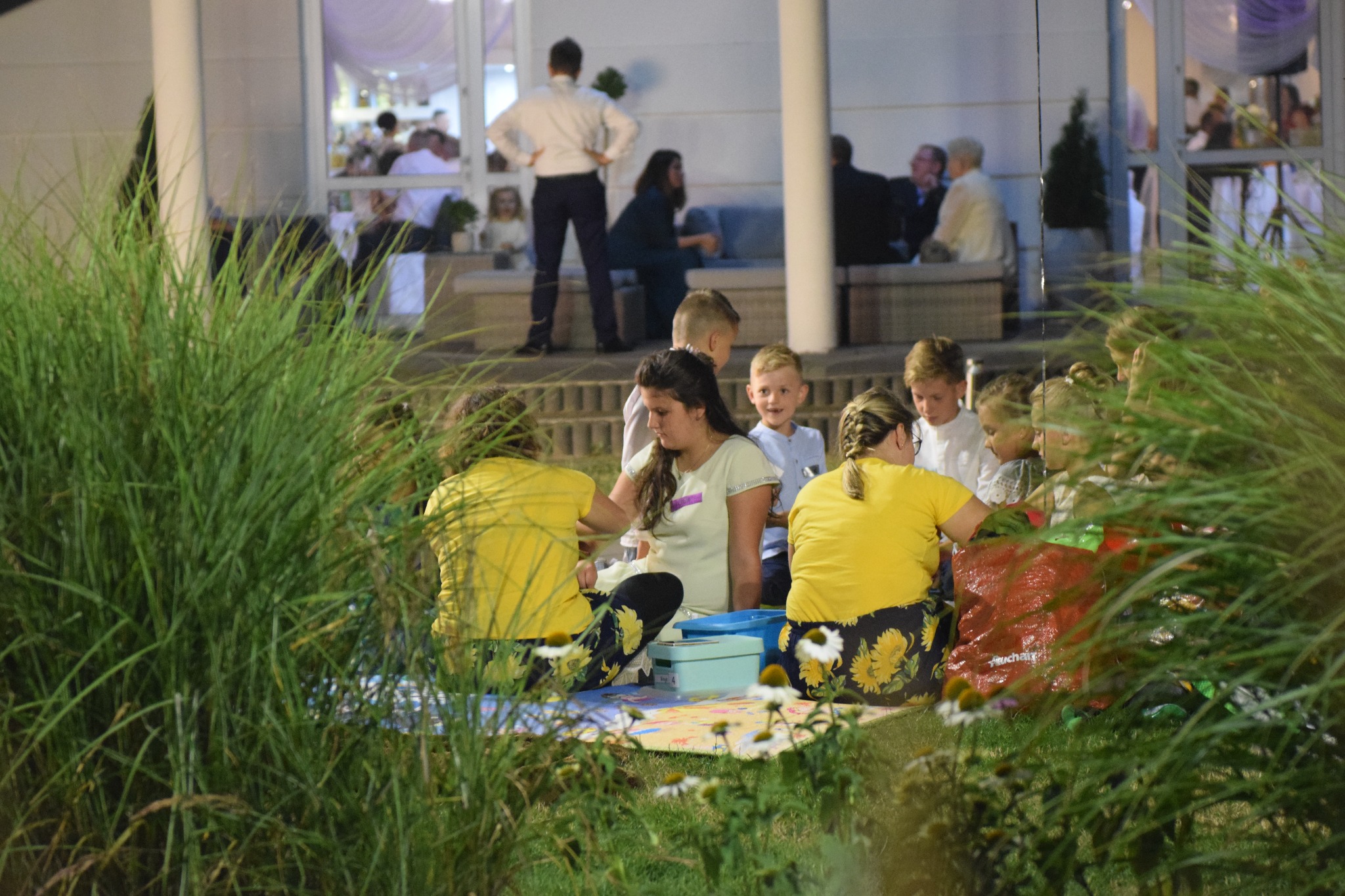 Dwie kobiety w żółtych koszulkach i dzieci siedzą na kocu na trawie, być może podczas animacji na świeżym powietrzu, w tle widać budynek i osoby.