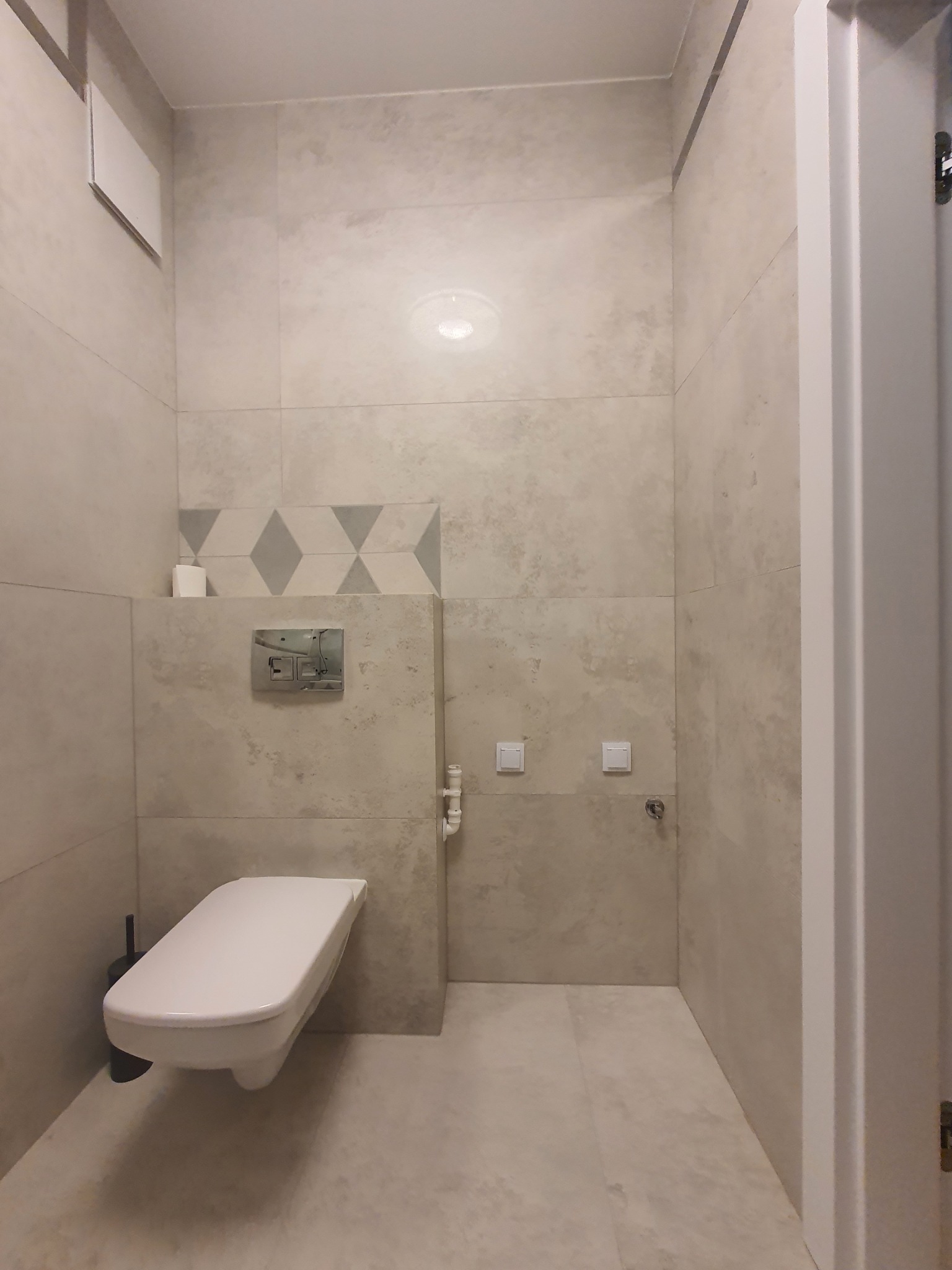 Nowoczesna łazienka z podwieszaną toaletą, płytkami imitującymi beton i dekoracyjnym pasem z rombami, oświetlona punktowo.