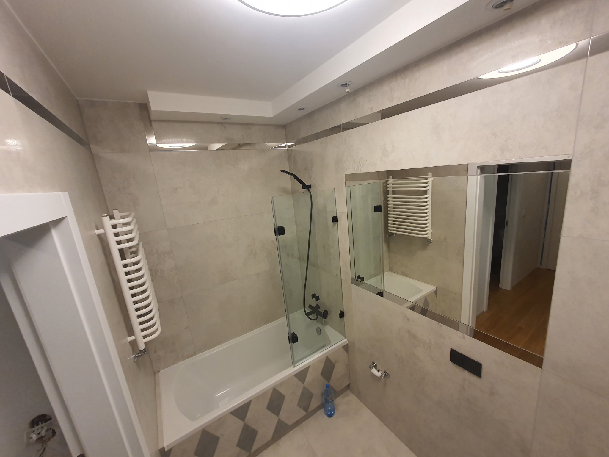 Nowoczesna łazienka z beżowymi płytkami, kabiną prysznicową z czarną armaturą i dużym lustrem, w którym odbija się fragment korytarza z drewnianą podłogą. Na podłodze stoi butelka wody mineralnej.