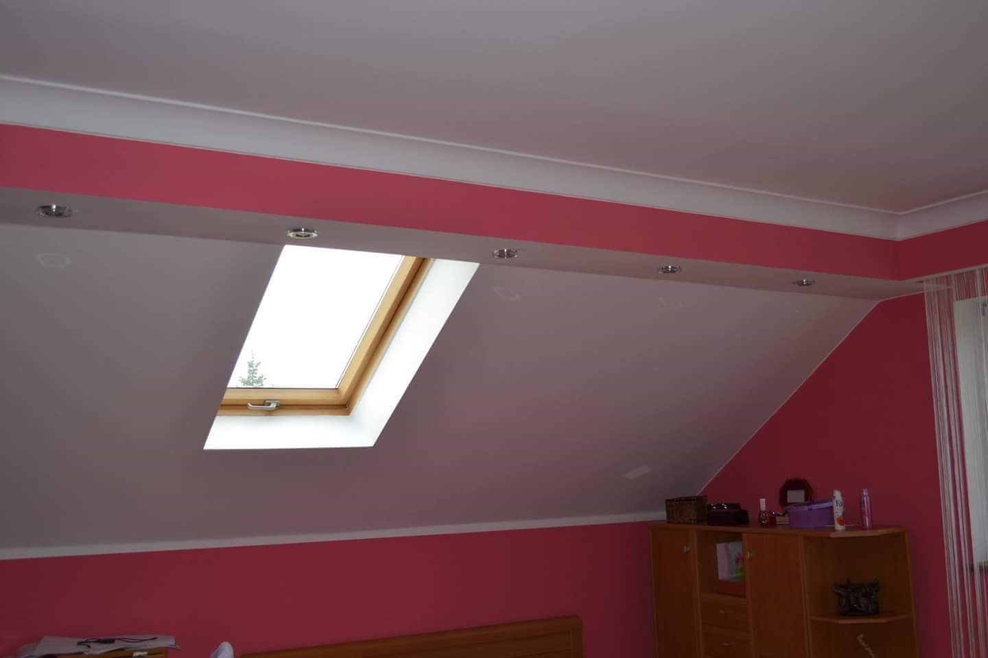 Pokój na poddaszu z oknem dachowym, białym sufitem z wpuszczonymi lampkami i różowymi ścianami. Widoczny fragment drewnianego łóżka i komody.