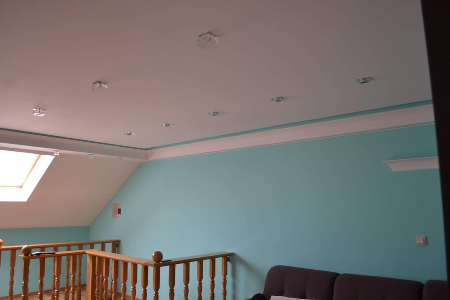 Poddasze z oknem dachowym, pomalowane na turkusowo ściany z białym gzymsem pod sufitem, biały sufit z zamontowanymi punktowymi lampami, drewniana balustrada schodów.