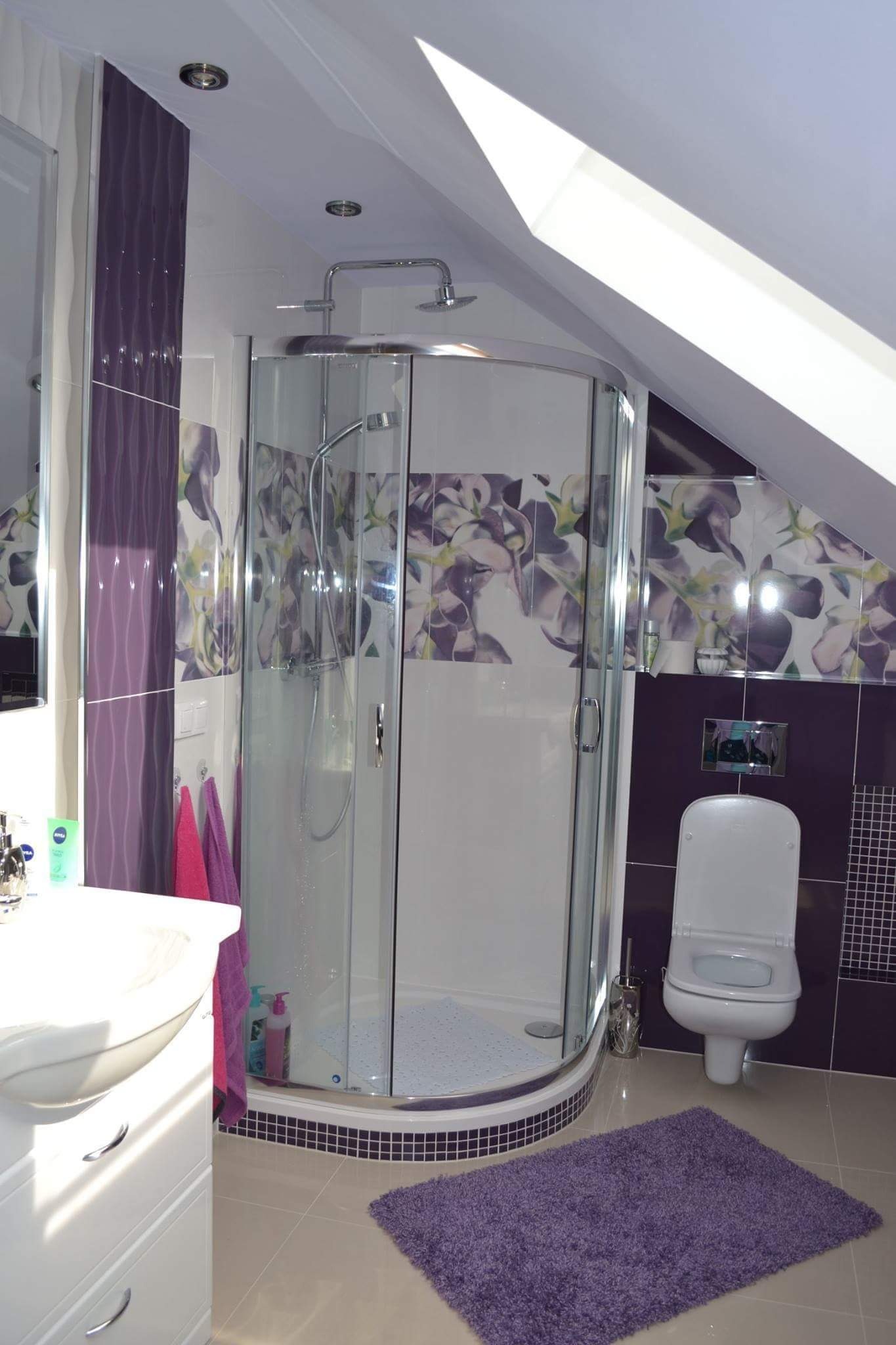 Jasna łazienka na poddaszu z fioletowymi akcentami: kabina prysznicowa z hydromasażem, umywalka z szafką, toaleta i fioletowy dywanik. Uwagę przykuwają płytki z motywem kwiatowym i dekoracyjne...